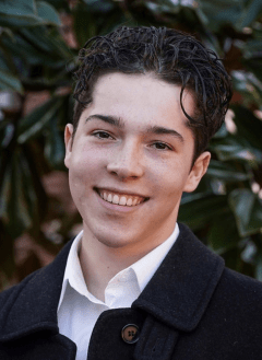 Close-up of Dominic Feder Di Toro, who has short curly dark hair and wears a white shirt and black coat.